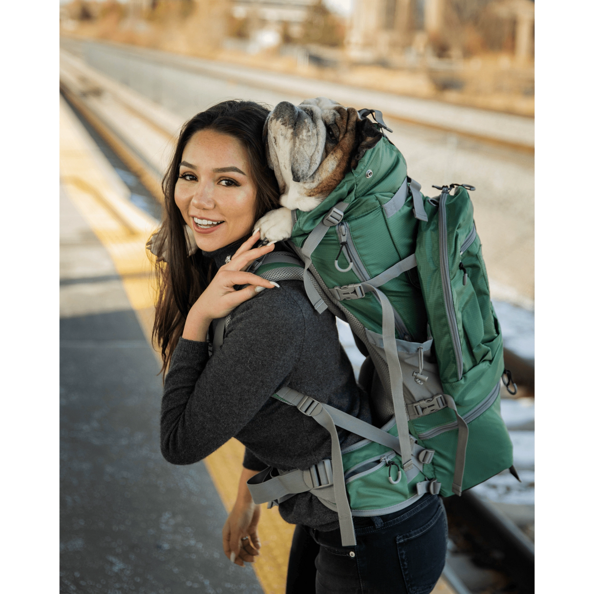 Woman carrying a bulldog in a backpack, smiling at a train station.